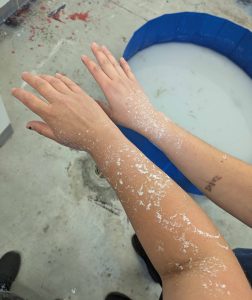 Profile of one of each of the artist's arms with a kiddie pool in the background on the floor. Their arms are covered in paper pulp.